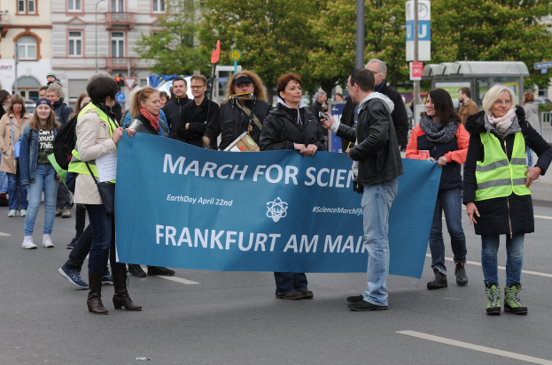 Gruppe von Menschen, die eine "March for Science Frankfurt am Main"-Tafel mit Bäumen, Pfählen, Schildern und Gebäuden im Hintergrund und einem klaren blauen Himmel tragen.