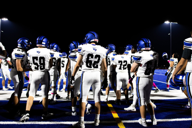 Gruppe von Füßballspielern in Helmen und Stiefeln auf einem Feld unter Flutlicht, bereit für ein Spiel.