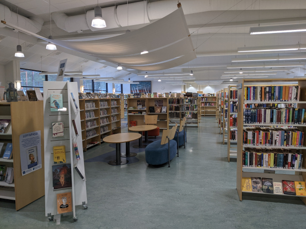 Eine geräumige Bibliothek mit Bücherregalen, Tischen, Stühlen, Sofas, Texttafeln, Beleuchtung und Rohren, mit Glasfenstern im Hintergrund.