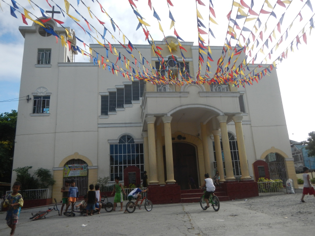 Eine Gruppe von Menschen, die auf Fahrrädern vor einer weißen Kirche mit architektonischen Details wie Fenstern, Säulen, Bögen, Türen und einem Kreuzsymbol fährt, vor einem bewölkten Himmel mit Pflanzen, einem Zaun, einer Fahne, Bäumen und bunten Fahnen, die an einem Seil befestigt sind.