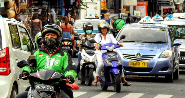 Eine Gruppe von Menschen auf Motorrädern auf einer belebten Straße mit Autos, Gebäuden, Bäumen und Geschäften im Hintergrund.