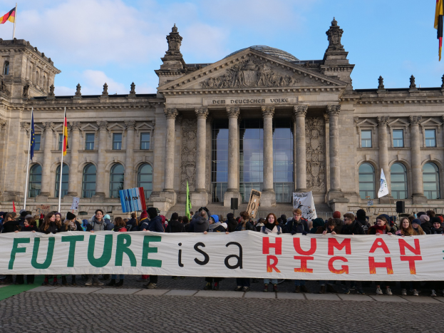 Eine Gruppe von Menschen mit einem Transparent mit der Aufschrift 'Zukunft ist ein Menschenrecht' vor dem Reichstagsgebäude in Berlin, Deutschland, mit sichtbaren Fenstern, Säulen und Bögen des Gebäudes und umgeben von Fahnen mit Stangen unter einem bewölkten Himmel.