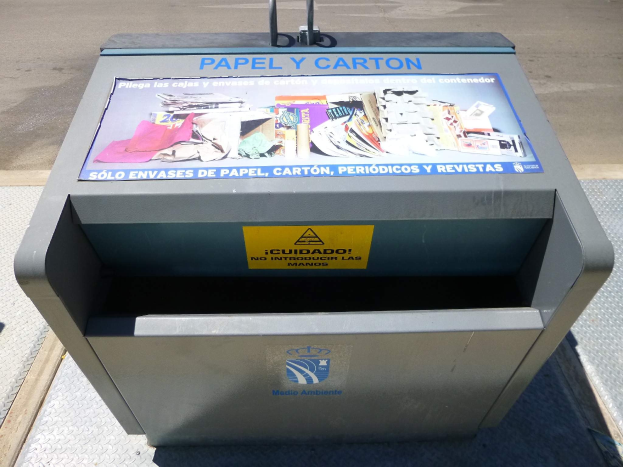 Metall-Mülltonne am Straßenrand mit einem "Papel y Carton"-Plakat, Fahrzeuge im Hintergrund sichtbar.