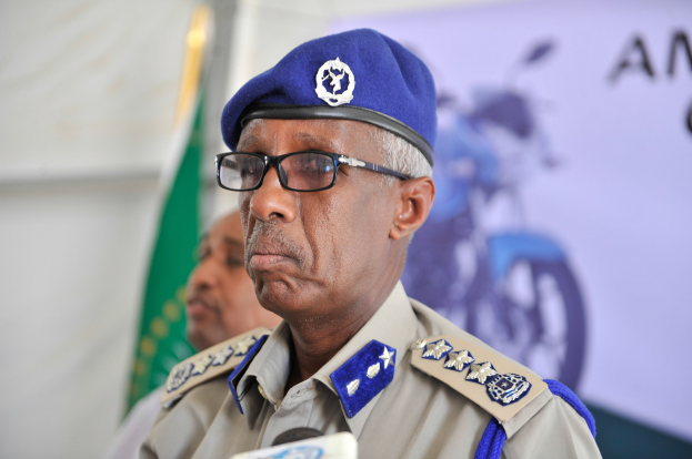 Ein Mann in Polizeiuniform mit Mütze und Brille steht an einem Pult mit Mikrofonen, neben ihm eine weitere Person, eine Fahne und ein Banner.