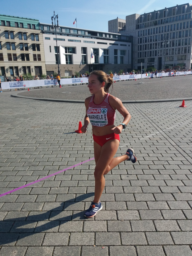 Eine Frau in Sportkleidung läuft bei einem Marathon, umgeben von Hütchen, einer Schleife, Zuschauern, einem Banner, Straßenlaternen mit Fahnen, Gebäuden und einem bewölkten Himmel.