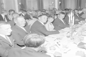 Eine Gruppe von Menschen sitzt um einen langen Tisch, lächelt und genießt die Gesellschaft des anderen, mit Gläsern, Besteck, Kerzen und anderen Gegenständen auf dem Tisch und Vorhängen und Wandlampen im Hintergrund, die eine gemütliche Atmosphäre schaffen.