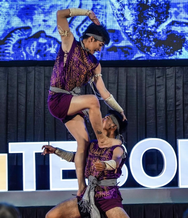 Zwei Performer in violetten Kleidern führen einen akrobatischen Hebemanöver auf der Bühne vor einem Publikum aus, mit einem schwarzen Vorhang und einer Leinwand im Hintergrund.