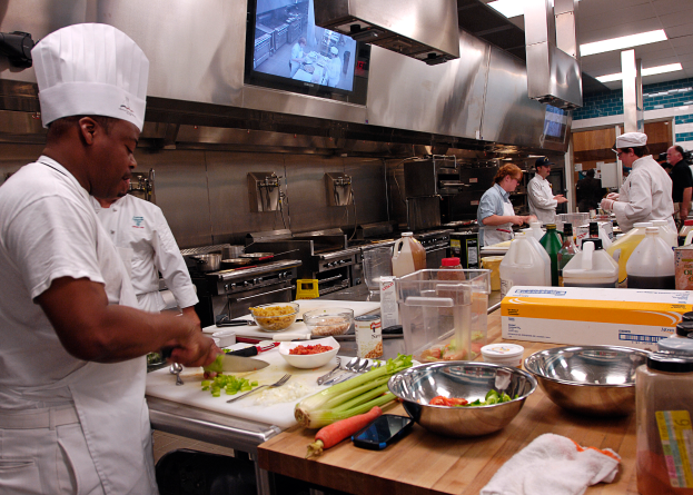Eine Gruppe von Köchen in einer Küche, die Essen auf einem Schneidbrett zubereiten, wobei ein Koch im Vordergrund einen weißen Dress und eine Haube trägt und ein Messer hält und von verschiedenen Utensilien, Schüsseln, Gemüse, einem Mobiltelefon, Flaschen und anderen Gegenständen umgeben ist, während im Hintergrund ein Fernseher zu sehen ist.