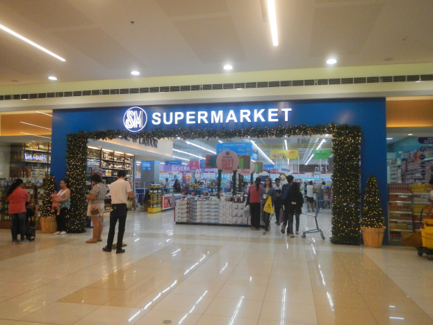 Gruppe von Menschen vor einem Supermarkt mit Einkaufstägern, einigen mit Einkaufstägern, einem Wagen, Topfpflanze, Regalen, Schildern und Deckenbeleuchtung.