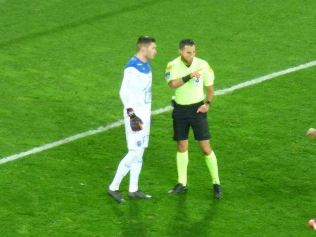 Ein Schiedsrichter in gelbem T-Shirt und schwarzen Shorts spricht mit einem Fußballspieler in weiß-blauer Uniform auf dem Feld, beide tragen Turnschuhe.