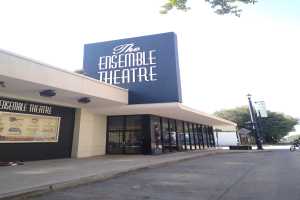 Außenansicht des Ensemble Theaters in San Francisco mit Glaswänden, einer Person auf dem Bürgersteig, einem Fahrrad auf der Straße, einem Schild, einem Straßenschild, Bäumen und einem bewölkten Himmel.