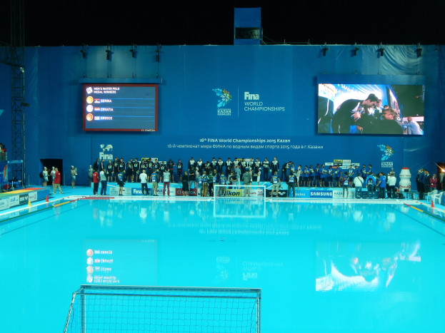 Eine Gruppe von Menschen, die um ein Hallenbad mit Netz am Boden stehen, mit einem Bildschirm im Hintergrund, auf dem "FINA World Championships 2015 Kazan" angezeigt wird.