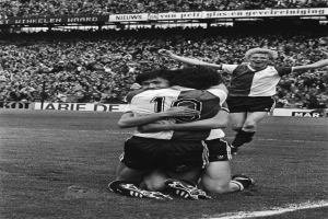 Drei Fussballspieler umarmen sich auf dem Boden und feiern ein Tor vor einer jubelnden Menge in einer schwarz-weißen Stadionszene.