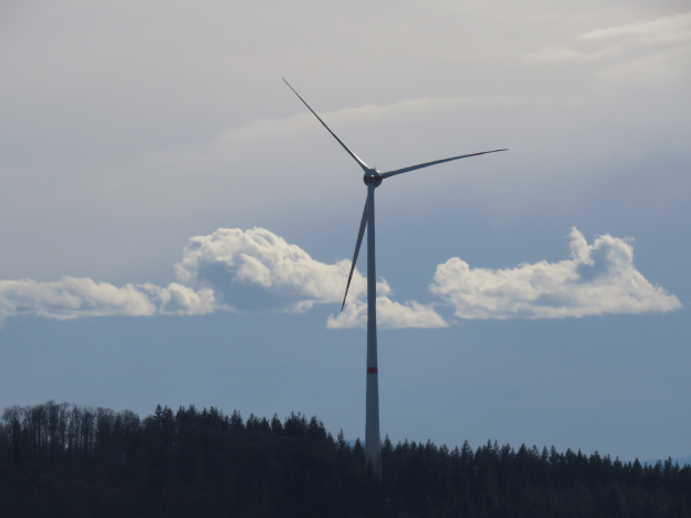 Windkraftanlage im Wald mit bewölktem Himmel und Bergen im Hintergrund.