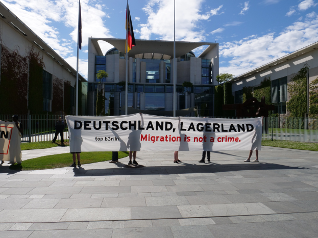 Gruppe von Menschen mit einem Banner "Deutschland, Lagerland Migration ist kein Verbrechen" vor einem Gebäude mit Fenstern, umgeben von Gras, einem Metallzaun, Pflanzen, Bäumen und Fahnenstangen.