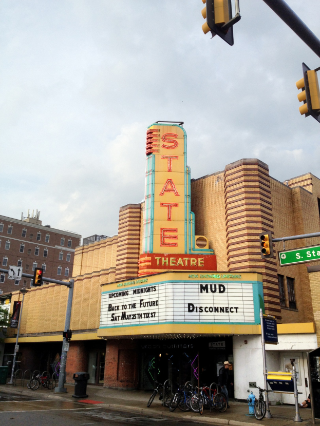 Außenansicht des Staatstheaters in Detroit, Michigan, mit einem Schild, parkenden Fahrrädern, einer Person und städtischen Elementen unter einem bewölkten Himmel.