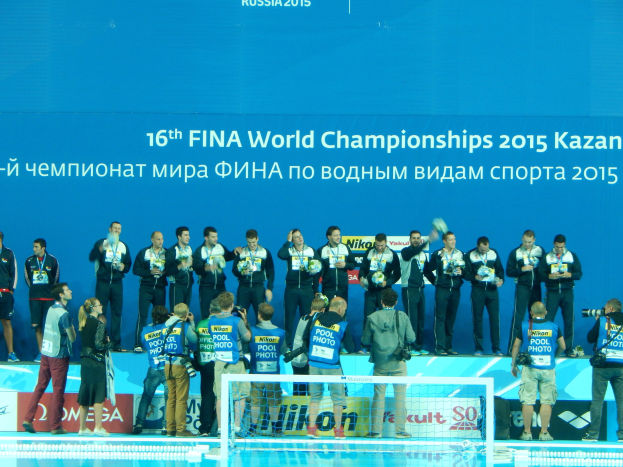 Gruppe von Menschen auf einem Schwimmbeckenrand mit Kameras, einem Netz im Vordergrund und einem "FINA World Championships 2015 Kazan"-Schild im Hintergrund.
