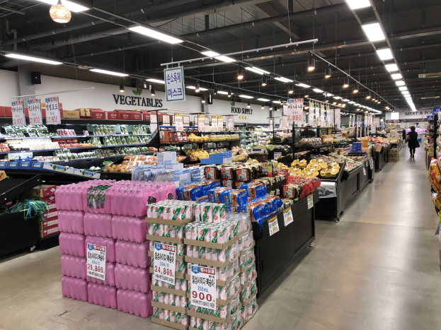 Ein hell erleuchteter japanischer Supermarktgang mit verschiedenen Lebensmitteln wie Obst und Gemüse, Deckenbeleuchtung, Informationsschildern und einigen Einkäufern.