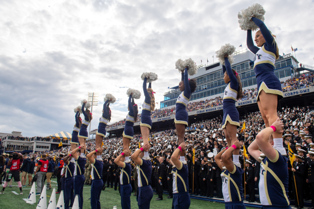 Eine Gruppe von Cheerleadern in blauen und weißen Uniformen führt einen Stunt auf einem Stadionevent aus, während sie Pompons halten und von einer Menge und einer Person gefilmt werden.