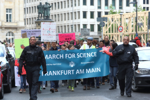 Eine Gruppe von Menschen marschiert mit einem "March for Science Frankfurt am Main"-Schild die Straße entlang, Autos, Gebäude, Statuen, Laternenmasten, Schilder und Bäume im Hintergrund.