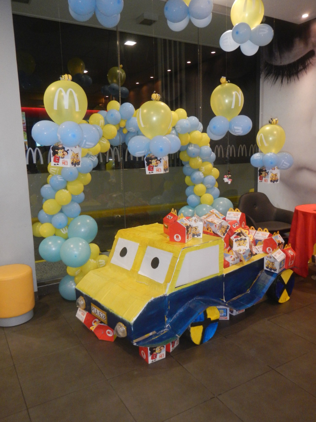 Eine festliche McDonald's-Geburtstagsparty-Szene mit einem gelben und blauen Lastwagen, der mit Ballons geschmückt ist, einem rot gedeckten Tisch mit Gegenständen, Hockern und einer Glaswand im Hintergrund unter Deckenlampen.