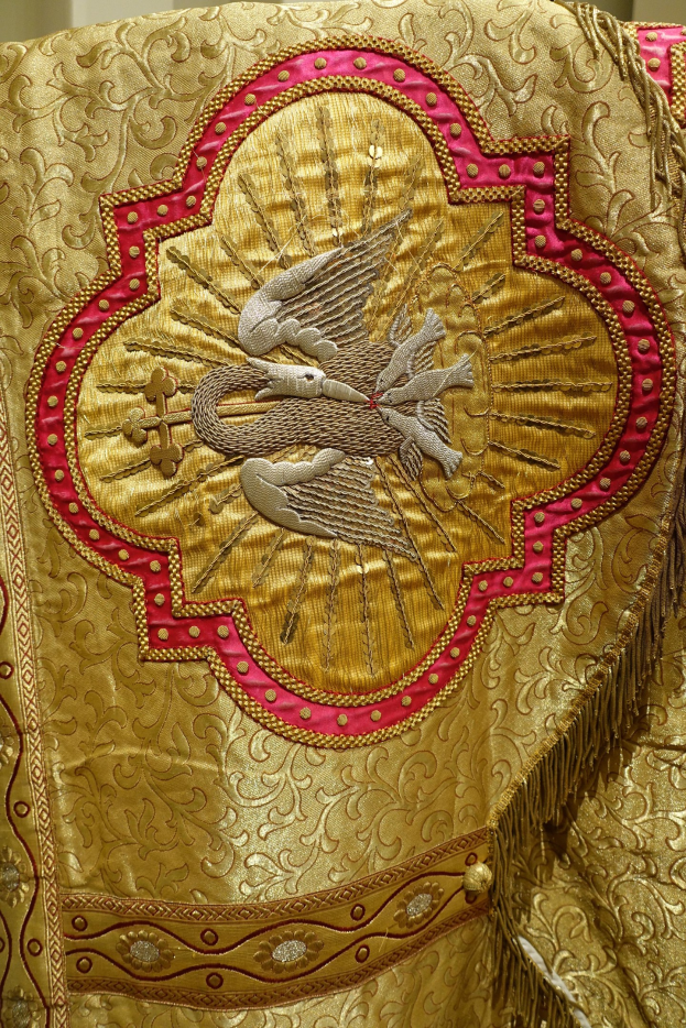 Nahaufnahme eines Priestergewandes mit aufwendiger goldener und roter Stickerei mit einem königlich anmutenden Design, das an einer Wand präsentiert wird.