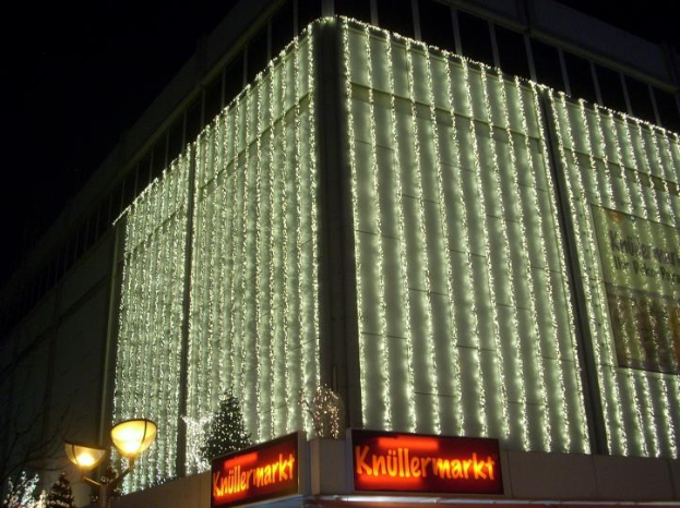 Ein Gebäude in Berlin, Deutschland, geschmückt mit Weihnachtslichtern, umgeben von Bäumen und Lichtmasten, mit Texttafeln in der Nähe und dem Himmel im Hintergrund.