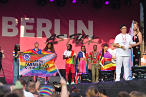 Gruppe von Menschen auf einer Bühne mit einem Banner, auf dem "Berlin as Fuck" steht, mit Lautsprechern, Mikrofonen und einer feiernden Menge unten.