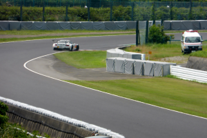 Ein Rennwagen fährt auf einer Rennstrecke mit einem Polizeiwagen im Hintergrund, umgeben von Gras, Pflanzen, Bäumen und einem Fahrzeug auf der rechten Seite und Text am unteren Rand.