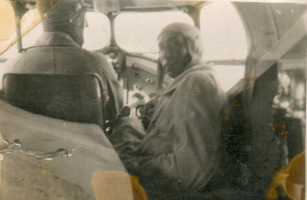 Schwarzes und weißes Foto von zwei Männern in einem Cockpit, einer sitzt im Pilotensitz und trägt eine Uniform, der andere steht neben ihm, beide haben ernste Gesichter.