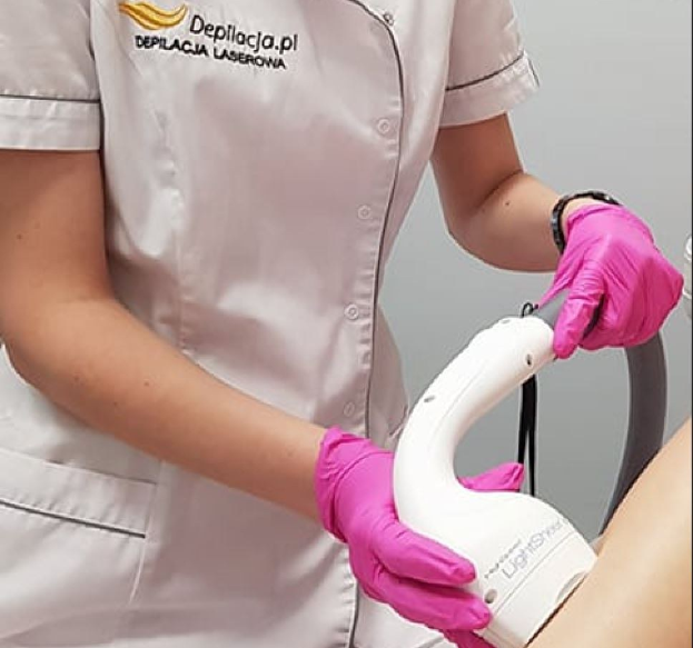 Eine Frau in einem weißen Hemd und pinken Handschuhen verwendet eine weiße Laser-Haarterminationsmaschine auf ihrer Haut, steht vor einer Wand.