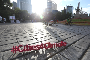 Gruppe von Menschen, die auf Fahrrädern eine Straße entlangfahren, mit dem Text "Cities4Climate" und im Hintergrund Gebäude, Bäume und andere städtische Elemente