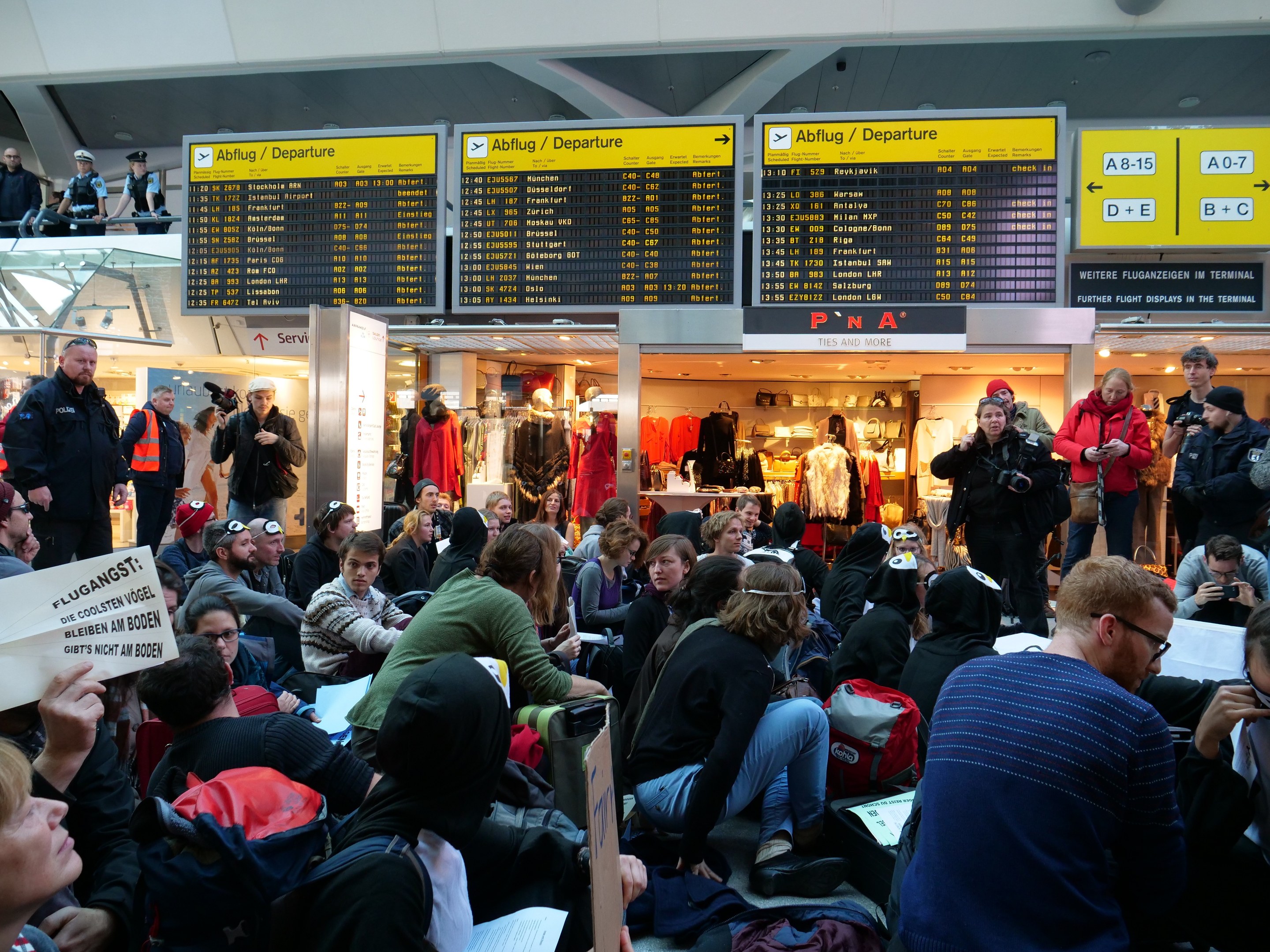 Eine große Gruppe von Menschen in einem Flughafen, einige sitzen mit Taschen und Papieren, andere stehen, mit Texttafeln, Schaufensterpuppen in Kleidern und Deckenleuchten im Hintergrund, was auf eine Demonstration hinweist.