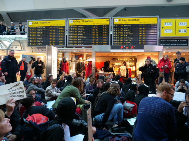 Eine große Gruppe von Menschen in einem Flughafen, einige sitzen mit Taschen und Papieren, andere stehen, mit Texttafeln, Schaufensterpuppen in Kleidern und Deckenleuchten im Hintergrund, was auf eine Demonstration hinweist.