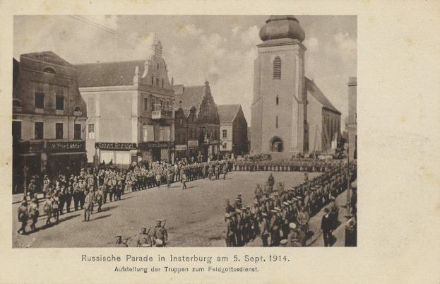 Ein Schwarz-Weiß-Foto eines Umzugs in Insterburg am 5. September 1914, das viele Menschen zeigt, einige stehen und einige gehen, mit Gebäuden im Hintergrund und Text unten auf dem Bild.