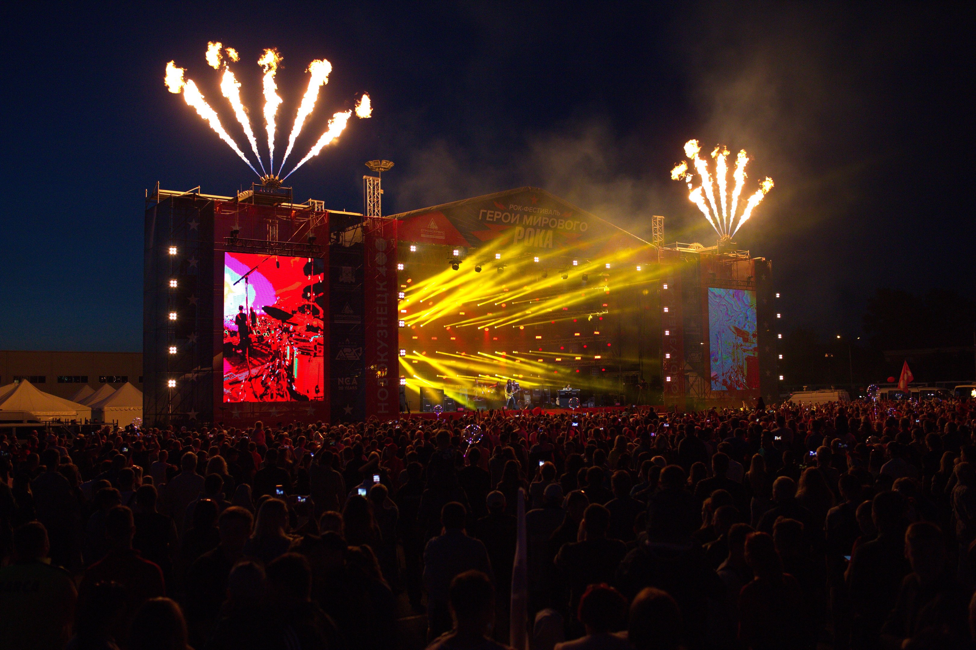 Große Menschenmenge auf einem Musikfestival mit beleuchteter Bühne, auf der Musiker spielen, umgeben von Bannern und Zelten links und Feuerwerk am dunklen Himmel.
