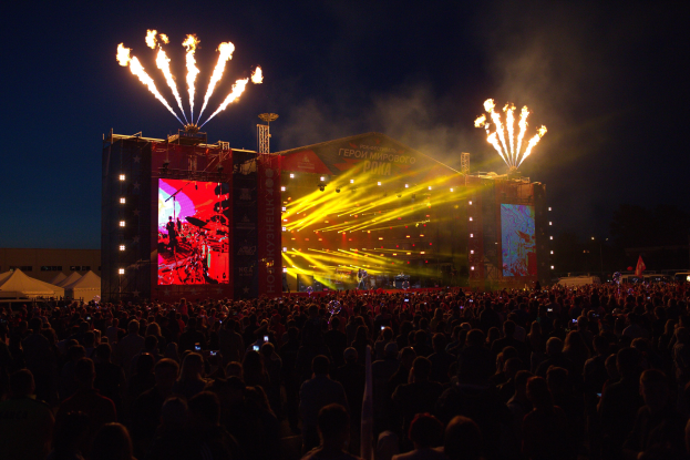 Große Menschenmenge auf einem Musikfestival mit beleuchteter Bühne, auf der Musiker spielen, umgeben von Bannern und Zelten links und Feuerwerk am dunklen Himmel.