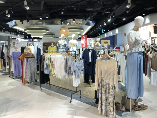 Ein hell erleuchteter Modegeschäft mit Kleiderstangen, Schaufensterpuppen in Outfits, Informationsschildern, Deckenleuchten und verstreuten Bodenartikeln.
