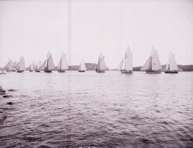 Schwarzes und weißes Foto von Segelbooten auf dem Wasser mit Hügeln im Hintergrund und einem klaren Himmel, beschriftet als Foto des New York Yacht Club.