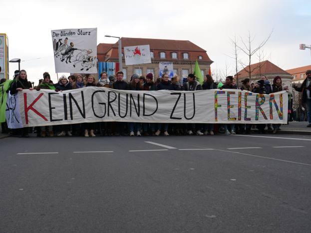 Demonstranten mit einem Banner mit der Aufschrift 'Kein Grund zu Feiern' gegen deutsche Sparmaßnahmen vor einem Gebäude.