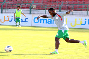 Ein Mann schießt einen Fußball auf einem Rasenfeld in einem Stadion, umgeben von zwei Personen und Bannern, Geländern und Stühlen.