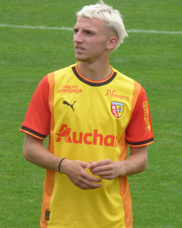 Ein Fußballspieler in einem gelb-orangen gestreiften Trikot steht selbstbewusst auf einem Feld und blickt entschlossen nach vorne.