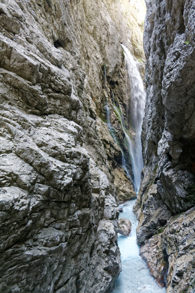 Ein kleiner Wasserfall ergießt sich über zerklüftete Felsen in einem steinigen Canyon, umgeben von saftig grünen Hügeln unter strahlendem Sonnenschein.