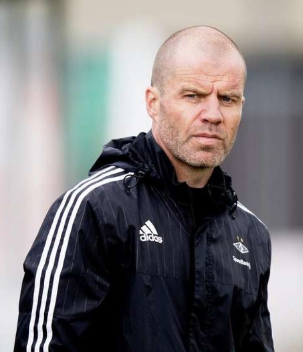 Kahlköpfiger Trainer in marineblauer Jacke mit weißen Streifen und weißem Kragen auf einem Fußballfeld stehend mit verschränkten Händen und ernster Miene.