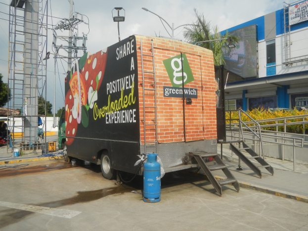 Ein großer Lastwagen parkt vor einem Gebäude mit einer Tafel, auf der "Teilen Sie eine positiv überladene Erfahrung" steht, mit Stufen, einem Zylinder am Boden, Geländern, Strommasten mit Drähten, Laternenmasten, Bäumen und einem bewölkten Himmel im Hintergrund.