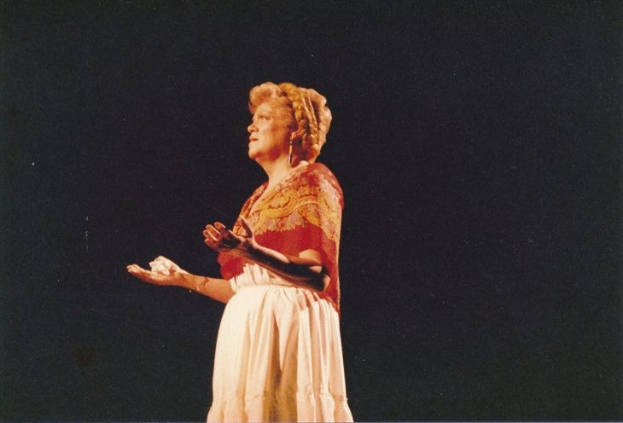 Eine Frau in einem weißen, hochgeschlossenen, langärmeligen Kleid steht auf einer dunklen Bühne mit ausgestreckten Händen, ihr Haar ist zurückgebunden und ihr Gesichtsausdruck ernst.