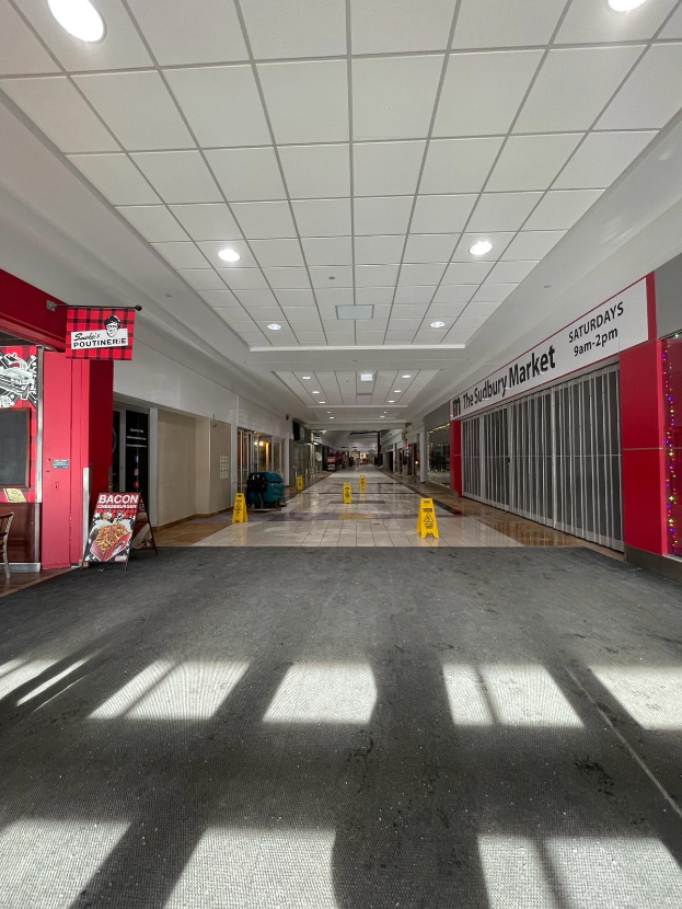 Hell erleuchtetes Einkaufszentrum-Innere mit einem Stuhl und einer Tafel, die "Sydbury Market - Vorher und Nachher" links zeigt, verstreute Gegenstände rechts und einen sichtbaren Boden.