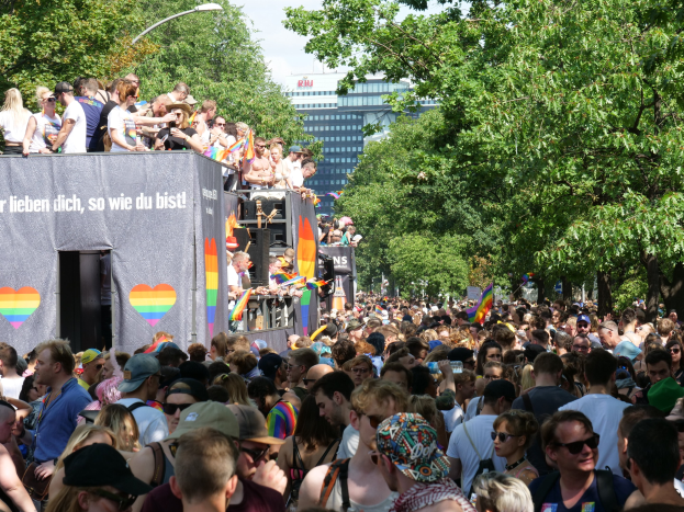 Eine große Menschenmenge steht vor einem Lastwagen bei der Christopher Street Day Parade in Berlin, viele tragen Mützen und Schutzbrillen, einige halten Fahnen, mit einem Banner auf dem Lastwagen und Bäumen, Gebäuden und einem Laternenpfahl im Hintergrund bei einem bewölkten Himmel.