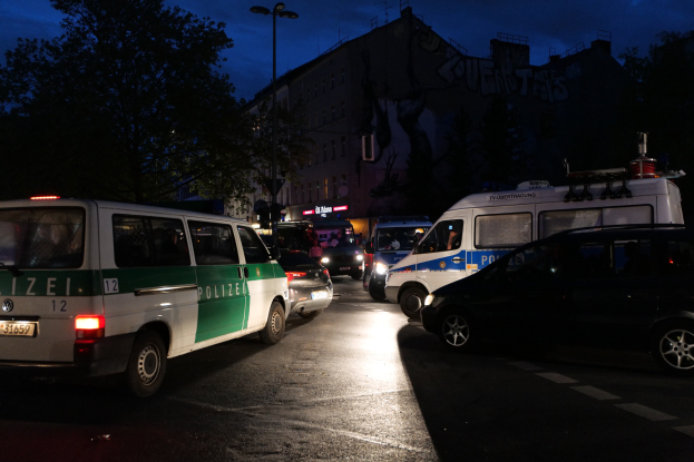 Eine Gruppe von Polizeiwagen, darunter ein Van, ein Auto und ein Rad, parkten nachts am Straßenrand, mit Gebäuden, Bäumen, Laternen und einem sternenklaren Himmel im Hintergrund.