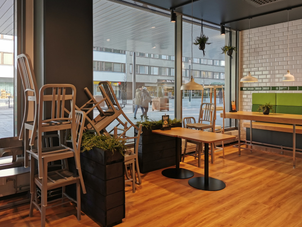 Ein Restaurant-Innenraum mit Holztischen und -stühlen, großen Fenstern, Pflanzen und Beleuchtung, mit Blick auf eine Berliner Straße mit Fußgängern, Gebäuden und Beschilderung.
